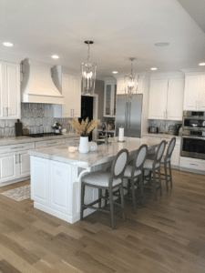 A bright, modern kitchen featuring white cabinets, a large central island with bar seating, pendant lighting, and light toned hardwood flooring.